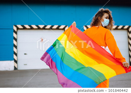 African American lesbian woman with medical face mask posing with lgbt pride flag. Curly woman during quarantine of coronavirus pandemic. African American lesbian woman with medical face mask posing with lgbt pride flag. Curly woman during quarantine of coronavirus pandemic. 101840442