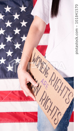 Woman is holding a sign Womens Rights Are Human Rights US flag on background. Protest against anti abortion law. Women's strike. Womens rights freedom. Woman is holding a sign Womens Rights Are Human Rights US flag on background. Protest against anti abortion law. Women's strike. Womens rights freedom. 101841570