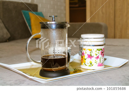 A glass coffee teapot and a ceramic sugar bowl stand on a tray on a gray sofa in the modern living room in a homely atmosphere. A cozy home at autumn season. Hot beverage to drink. Good morning. A glass coffee teapot and a ceramic sugar bowl stand on a tray on a gray sofa in the modern living room in a homely atmosphere. A cozy home at autumn season. Hot beverage to drink. Good morning. 101855686