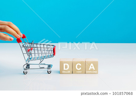The text DCA is written on wooden cubes and woman's hand pushingn empty shopping cart in studio on blue background. Business concept. DCA - short for dollar cost averaging. 101857068