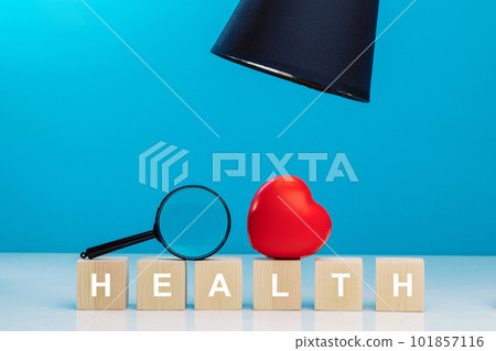 Red heart and magnifying glass on top of wooden cubes with the word HEALTH on a table next to a black table lamp. 101857116