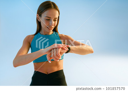 Smiling sporty woman standing and looking at watch checking her performance on studio background Smiling sporty woman standing and looking at watch checking her performance on studio background 101857570