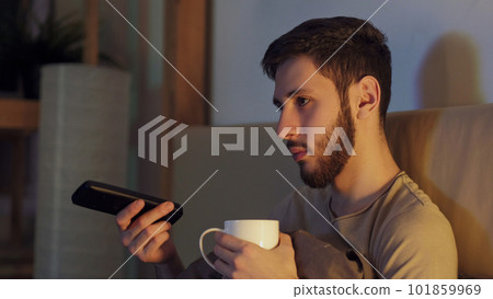 TV evening. Night insomnia. Home leisure. Tired bored guy with mug watching news switching channels with remote control drinking tea alone in dark room with blue light. TV evening. Night insomnia. Home leisure. Tired bored guy with mug watching news switching channels with remote control drinking tea alone in dark room with blue light. 101859969