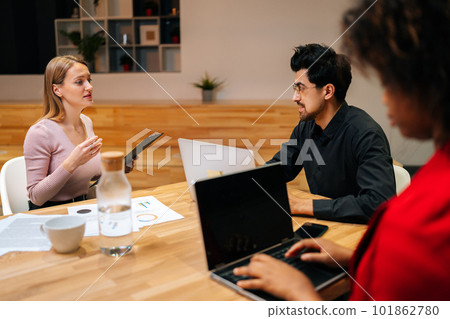 Remote view of elegant male team leader sitting at table with multiethnic colleagues, discussing working issues at negotiations meeting. Serious male businessman talking at diverse group meeting. Remote view of elegant male team leader sitting at table with multiethnic colleagues, discussing working issues at negotiations meeting. Serious male businessman talking at diverse group meeting. 101862780