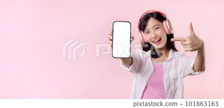 Happy cheerful smiling asian woman with wireless earphones showing blank screen mobile phone or new smartphone music application advertisement mockup isolated on pink studio background. Happy cheerful smiling asian woman with wireless earphones showing blank screen mobile phone or new smartphone music application advertisement mockup isolated on pink studio background. 101863163