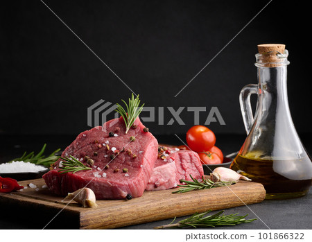 Raw piece of beef with spices pepper, rosemary sprig, salt and olive oil on a wooden board, black background Raw piece of beef with spices pepper, rosemary sprig, salt and olive oil on a wooden board, black background 101866322