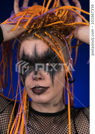 Fine art portrait of young woman with horror black stage make up painted on face and orange color dreadlocks hairstyle. Front view, studio shot on blue background. Part of photo series Fine art portrait of young woman with horror black stage make up painted on face and orange color dreadlocks hairstyle. Front view, studio shot on blue background. Part of photo series 101867356