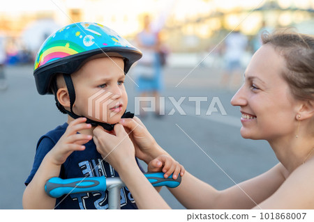 Close-up mom parent hand put on and fasten safety helmet on cute blond caucasian toddler boy for riding bike or scooter city street park outdoors on summer day. Child sport activity protection care 101868007
