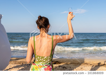 Back view portrait young adult woman holding cold alcohol refresh beverage glass looking at ocean shore at summer seaside vacation. Female person treat relax holiday at exotic resort hotel beach 101868009