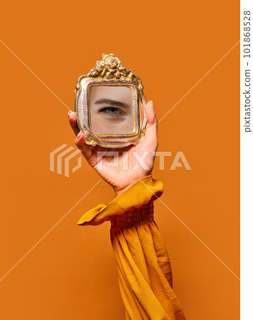 Elegant female hand holding small mirror with reflection of girl's eye with makeup isolated over orange background. Eyelash extension Elegant female hand holding small mirror with reflection of girl's eye with makeup isolated over orange background. Eyelash extension 101868528