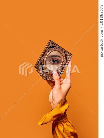Female hand holding small mirror with reflection of beautiful, green girl's eye over orange background. Eyelash lamination 101868578