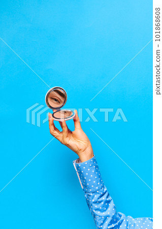 Graceful female hand holding small mirror with reflection of beautiful girl's eye glowing with joy over bright blue background Graceful female hand holding small mirror with reflection of beautiful girl's eye glowing with joy over bright blue background 101868608