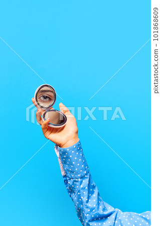 Graceful female hand holding small mirror with reflection of beautiful girl's eye with makeup isolated on blue background Graceful female hand holding small mirror with reflection of beautiful girl's eye with makeup isolated on blue background 101868609