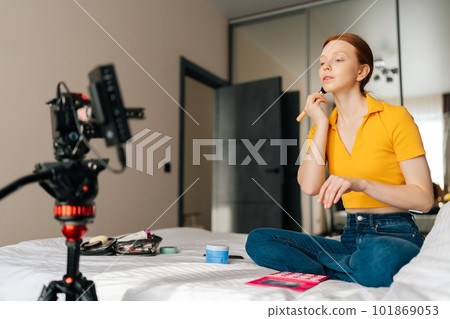 Low-angle view of redhaired female beauty blogger making video tutorial using professional camera standing on tripod, talking how to put makeup on step by step to include it on blog, sitting on bed. 101869053