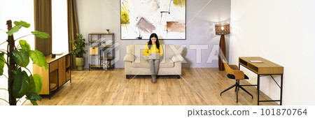 Young woman is working on laptop while sitting on couch in her comfortable living room. She is engaged in digital work, enjoying benefits of remote work and modern technology. High quality photo 101870764