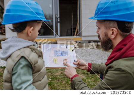 Father and his little son in front of their unfinished house holding picture with paper house with solar panels. Father and his little son in front of their unfinished house holding picture with paper house with solar panels. 101873568