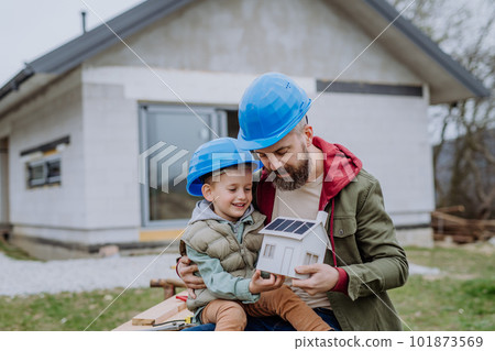Father and his little son in front of their unfinished house holding model of paper house with solar panels. Father and his little son in front of their unfinished house holding model of paper house with solar panels. 101873569