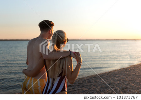 Rear view of young couple in striped swimsuits walking in the beach during sunset. 101873767