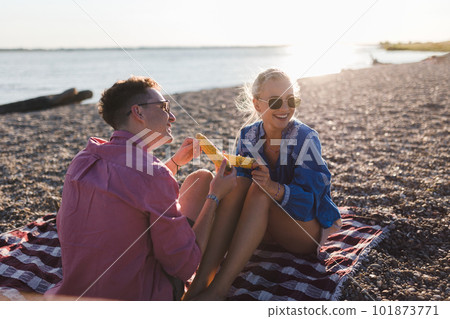 Happy young couple dating together in beach, sitting on a blanket and eating corn. Enjoying holiday time together. 101873771