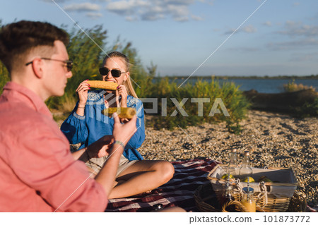 Happy young couple dating together in beach, sitting on a blanket and eating corn. Enjoying holiday time together. 101873772