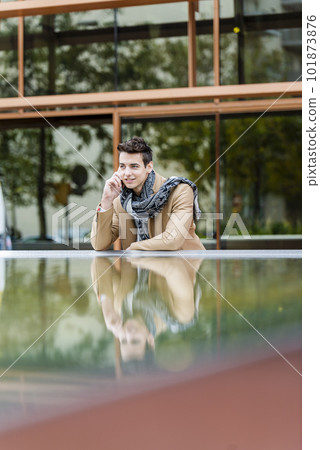 Portrait of stylish handsome young man with coat standing outdoors and leaning on wall using a smartphone 101873876