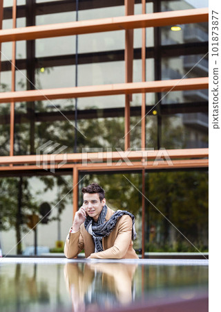 Portrait of stylish handsome young man with coat standing outdoors and leaning on wall using a smartphone 101873877