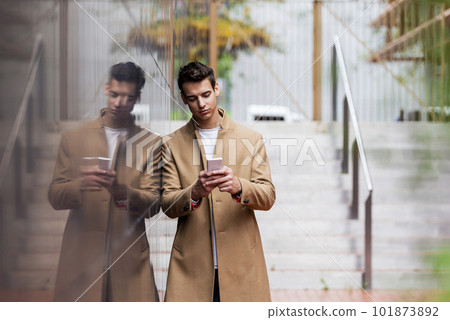 Portrait of stylish handsome young man with coat standing outdoors and leaning on wall using a smartphone 101873892