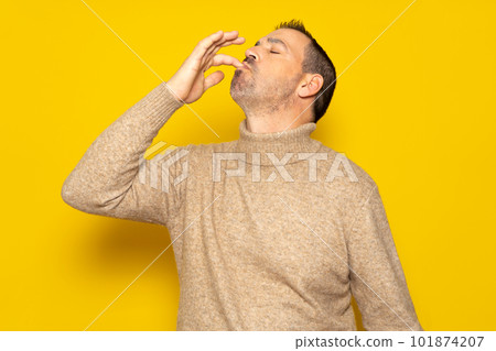 Bearded Hispanic man wearing a beige turtleneck smacking his lips and licking his fingers after devouring an exquisite delicacy, isolated on yellow studio background. 101874207