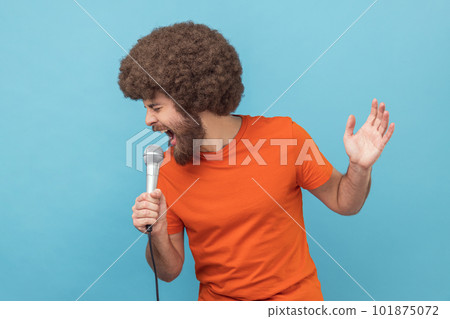 Portrait of man with Afro hairstyle wearing orange T-shirt singing songs, holding microphone, singer making performance, having fun on party in karaoke. Indoor studio shot isolated on blue background. 101875072