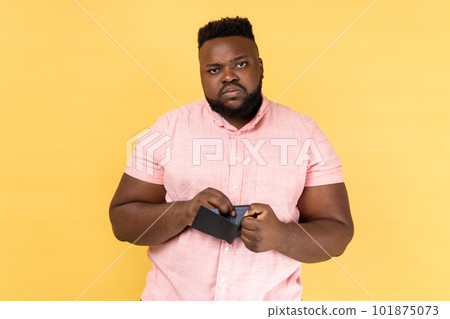 Portrait of sad poor depressed bearded man wearing pink shirt standing looking at camera with upset facial expression, showing empty wallet. Indoor studio shot isolated on yellow background. 101875073