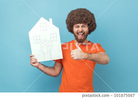Portrait of excited positive man with Afro hairstyle wearing orange T-shirt holding paper house, real estate agency, showing thumb up. Indoor studio shot isolated on blue background. 101875087