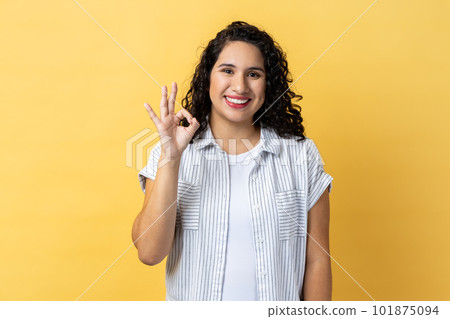 Portrait of smiling satisfied positive optimistic beautiful woman with dark wavy hair looking at camera showing ok sign gesture. Indoor studio shot isolated on yellow background. Portrait of smiling satisfied positive optimistic beautiful woman with dark wavy hair looking at camera showing ok sign gesture. Indoor studio shot isolated on yellow background. 101875094
