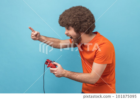 Side view of excited man with Afro hairstyle in orange T-shirt holding in hands red gamepad joystick, grimacing playing video games, raised finger up. Indoor studio shot isolated on blue background. Side view of excited man with Afro hairstyle in orange T-shirt holding in hands red gamepad joystick, grimacing playing video games, raised finger up. Indoor studio shot isolated on blue background. 101875095