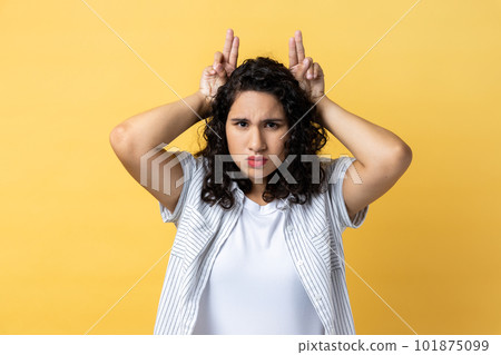 Portrait of strict serious woman with dark wavy hair showing bull horn gesture with fingers over head, looking hostile and threatening. Indoor studio shot isolated on yellow background. Portrait of strict serious woman with dark wavy hair showing bull horn gesture with fingers over head, looking hostile and threatening. Indoor studio shot isolated on yellow background. 101875099