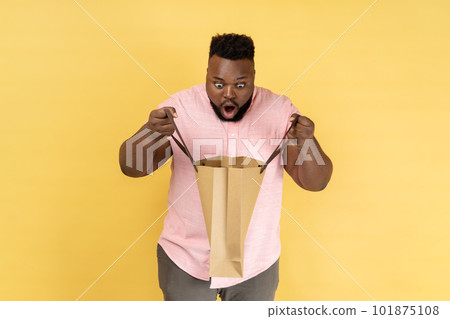 Portrait of man wearing pink shirt holding looking inside shopping bag, being astonished with cheap purchase or excellent gift. Indoor studio shot isolated on yellow background. 101875108