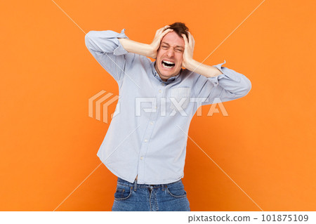 Portrait of upset sad young adult man standing with hands on head, suffering terrible headache, screaming from pain, wearing light blue shirt. Indoor studio shot isolated on orange background. 101875109