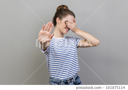 Portrait of afraid beautiful woman wearing striped T-shirt closing eyes with palm and showing stop hand gesture, turning face, does not want to see. Indoor studio shot isolated on gray background. 101875114