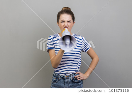 Portrait of woman in striped T-shirt holding megaphone near mouth loudly speaking, screaming, making announcement, paying attention at social problems. Indoor studio shot isolated on gray background. 101875116