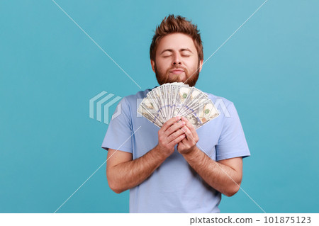 Portrait of satisfied bearded man smelling earned dollar banknotes, enjoying success and big profit, wealthy life, greedy for money. Indoor studio shot isolated on blue background. Portrait of satisfied bearded man smelling earned dollar banknotes, enjoying success and big profit, wealthy life, greedy for money. Indoor studio shot isolated on blue background. 101875123
