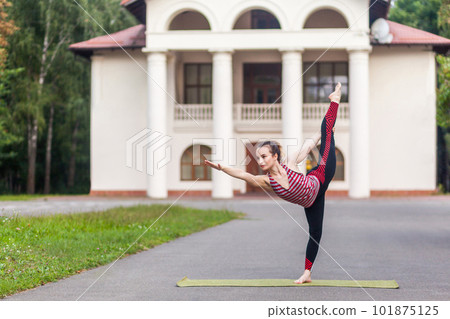 Full length portrait of positive caucasian woman in active wear enjoying yoga outdoor, balancing and feeling body recreation in morning, standing in asana on one leg, recreating keeping fit. 101875125