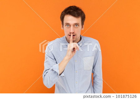 Serious young adult man standing with finger near lips, asking to keep silent, looking at camera with bossy expression, wearing light blue shirt. Indoor studio shot isolated on orange background. 101875126