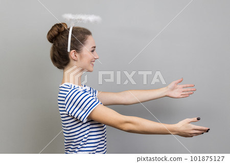 Side view of woman wearing striped T-shirt and with nimbus over her head stretching arms with kind friendly smile, going to embrace, share love. Indoor studio shot isolated on gray background 101875127