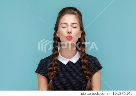 Portrait of romantic flirting woman with braids standing with closed eyes and pout lips, sending air kisses to her boyfriend, wearing black dress. woman Indoor studio shot isolated on blue background. 101875143
