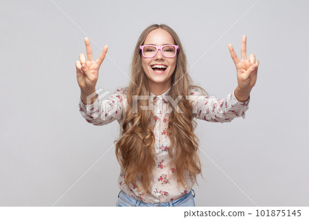 Belief in winning. Smiling pretty woman in glasses with wavy blond hair showing v sign with fingers, peace or victory gesture, rejoicing success. Indoor studio shot isolated on gray background. Belief in winning. Smiling pretty woman in glasses with wavy blond hair showing v sign with fingers, peace or victory gesture, rejoicing success. Indoor studio shot isolated on gray background. 101875145