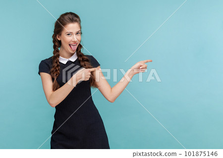 Portrait of cheerful lovely woman with braids sticks tongue, , indicates with both fore fingers at blank space, wearing black dress. woman Indoor studio shot isolated on blue background. Portrait of cheerful lovely woman with braids sticks tongue, , indicates with both fore fingers at blank space, wearing black dress. woman Indoor studio shot isolated on blue background. 101875146