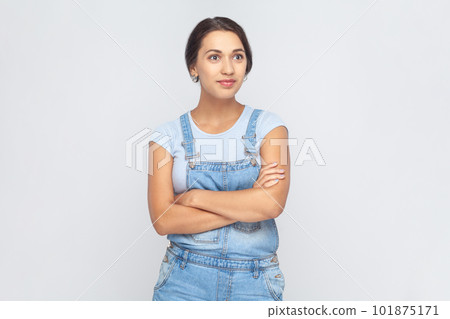 Portrait of calm smiling woman wearing denim overalls standing with crossed arms, looking at camera with confident facial expression. Indoor studio shot isolated on gray background. Portrait of calm smiling woman wearing denim overalls standing with crossed arms, looking at camera with confident facial expression. Indoor studio shot isolated on gray background. 101875171