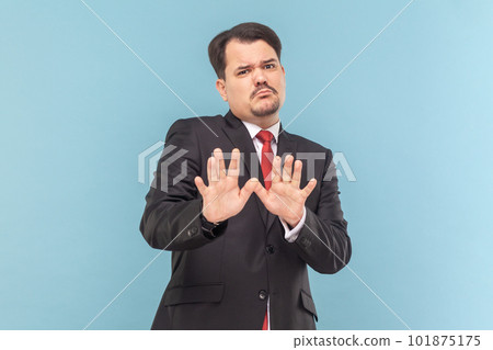 Portrait of frighten man with mustache standing raised arms, showing stop block gesture, being scared, wearing black suit with red tie. Indoor studio shot isolated on light blue background. Portrait of frighten man with mustache standing raised arms, showing stop block gesture, being scared, wearing black suit with red tie. Indoor studio shot isolated on light blue background. 101875175