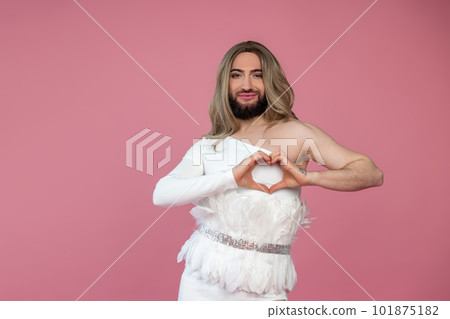 Smiling transgender wearing blonde wig and white dress making heart shape with hands expressing love 101875182