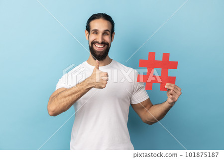 Portrait of happy man with beard wearing white T-shirt holding hashtag symbol, showing thumb up, recommending popular topics, internet trends. Indoor studio shot isolated on blue background. Portrait of happy man with beard wearing white T-shirt holding hashtag symbol, showing thumb up, recommending popular topics, internet trends. Indoor studio shot isolated on blue background. 101875187