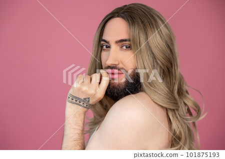 Transgender wearing blonde wig posing with bare shoulder looking at camera keeps finger near lips 101875193